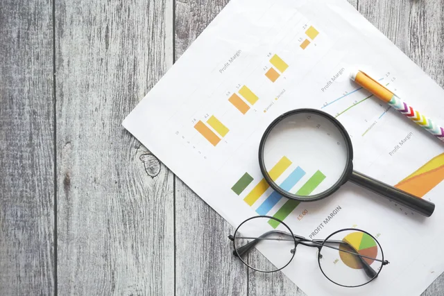 Overhead view of financial charts, a magnifying glass, ballpen, and eyeglasses on a wooden surface. Company descriptions: reading yours as a lender, then as an investor